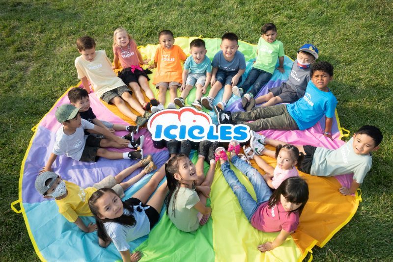 Kids in Icicles t-shirts sitting in a circle on a rainbow parachute around the Icicles logo